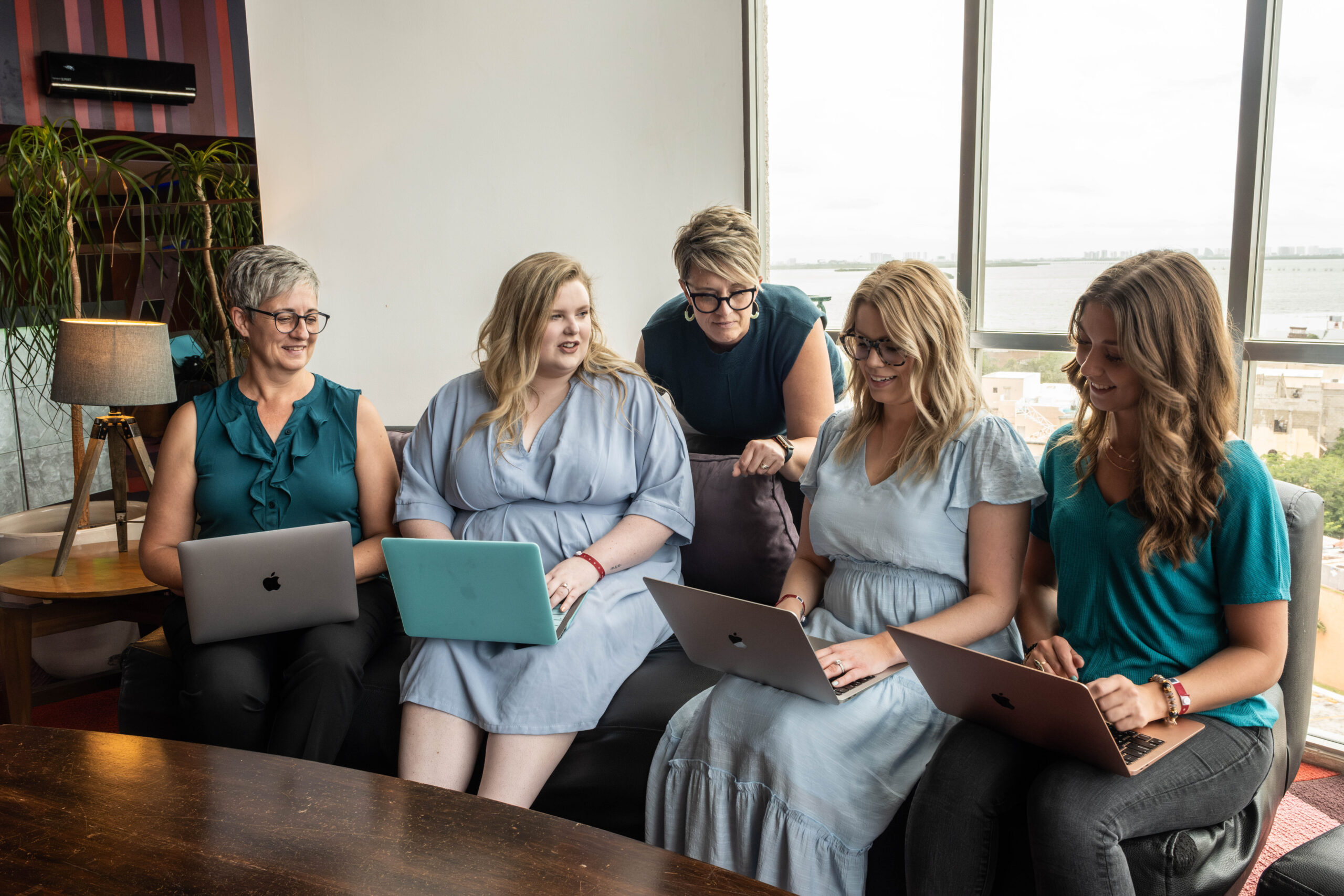 Team of five women collaborating on laptops in a bright office, reviewing work together—illustrating a hands-on approach to evaluating and improving business processes.